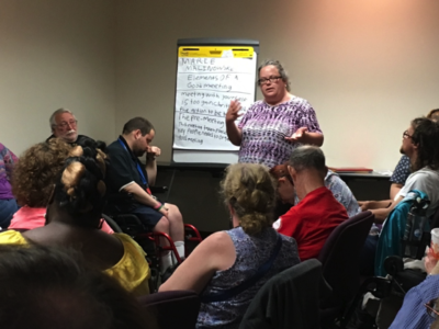Picture of Marie Malinowski, a woman, standing in front of a flip chart with topics written on it, facing an audience of self-advocates who are listening to her present.