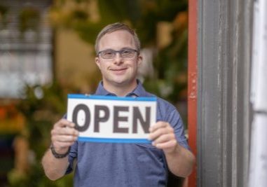 A man with Down Syndrome stands at the entrance to his shop as a proud owner and holds an open sign.