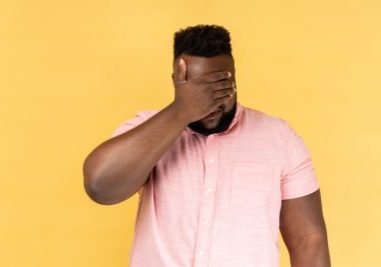 Portrait of man wearing pink shirt closing eyes with hand, dont want to see that, ignoring problems, hiding from stressful situations. Indoor studio shot isolated on yellow background.